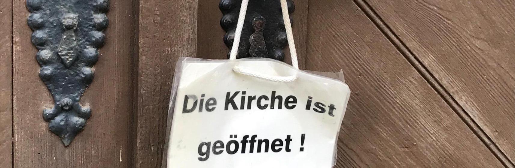 An der Türklinke einer Kirchentür hängt der Hinweis "Die Kirche ist geöffnet"